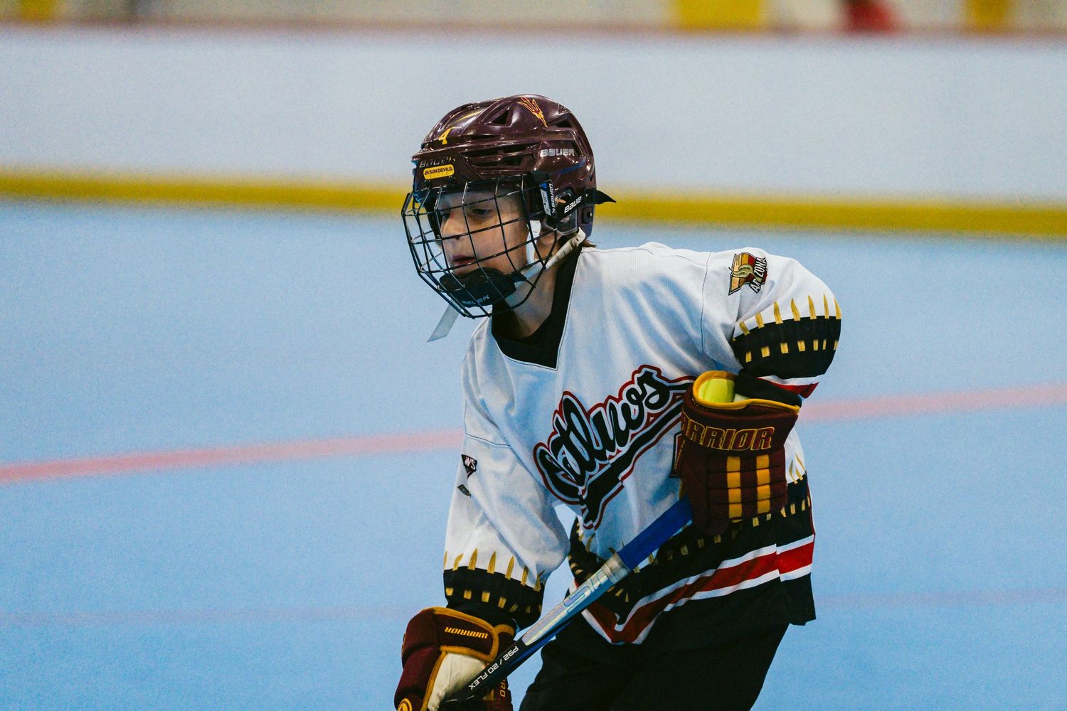 Young hockey player in white jersey holds stick on indoor rink, wearing helmet and gloves during a game.