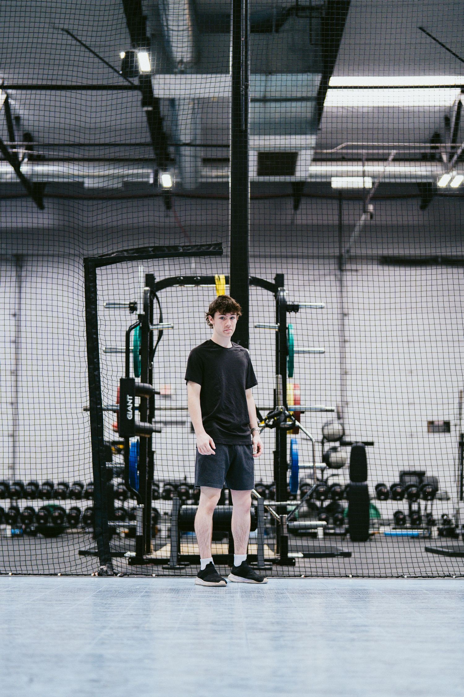 Teen athlete in black t-shirt and shorts stands in a modern gym with weight racks and netting.