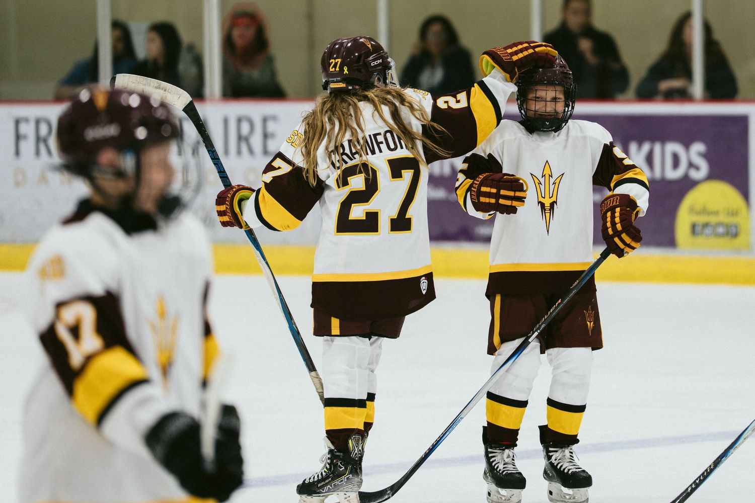 Arizona State Sun Devils women's hockey players celebrate on the ice during a game, wearing maroon and gold uniforms.