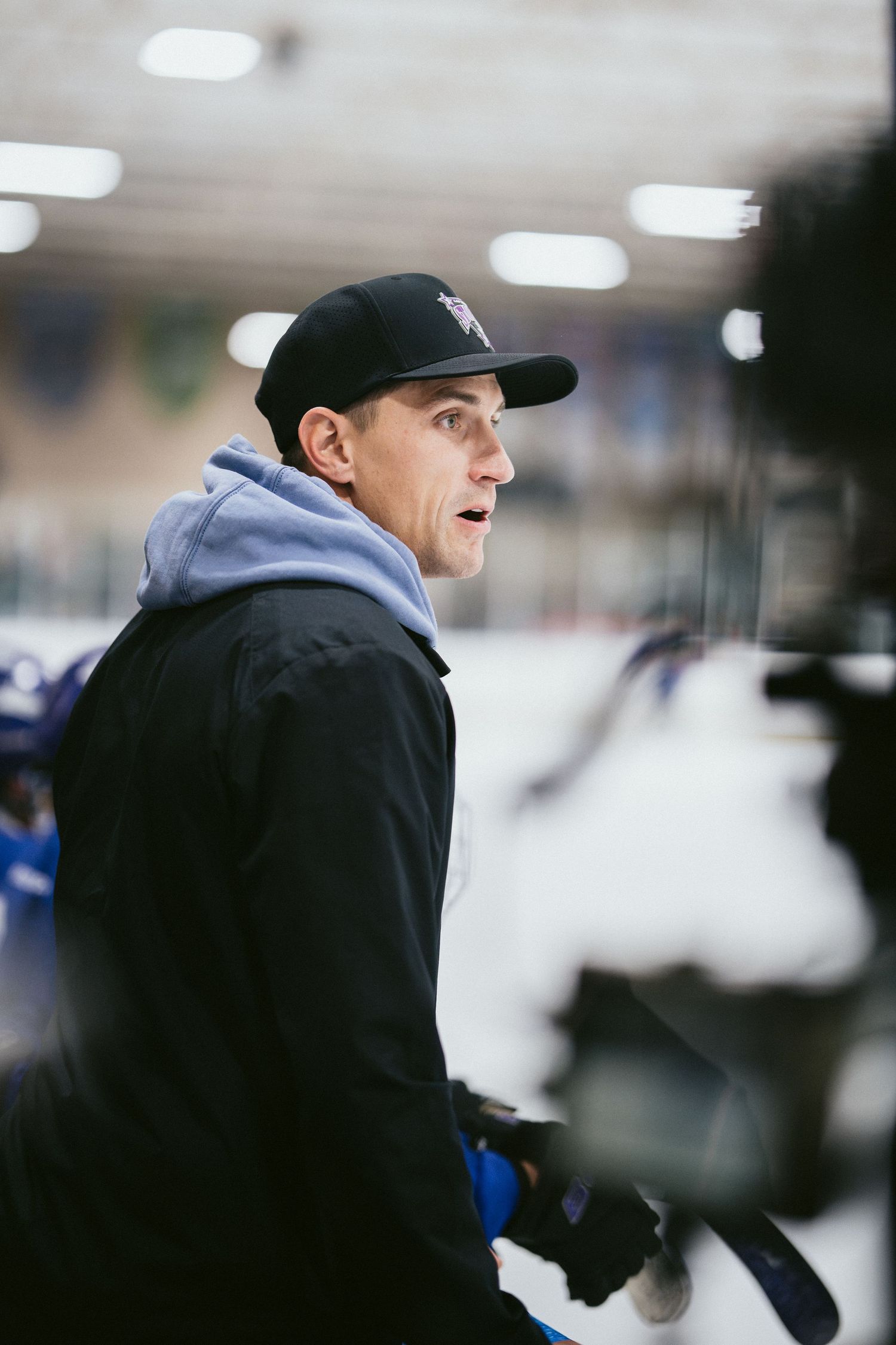 A hockey coach in a black jacket and cap stands rinkside, holding a stick during an indoor ice hockey practice.