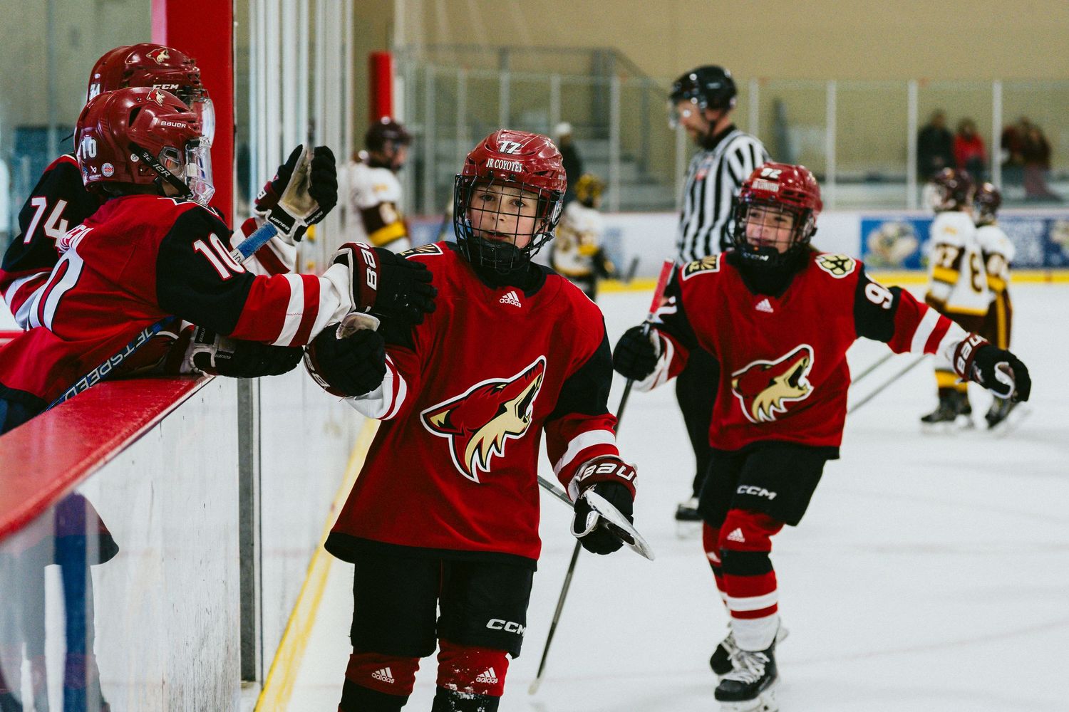 Youth hockey players in red and black jerseys skate during a competitive game on an indoor ice rink.
