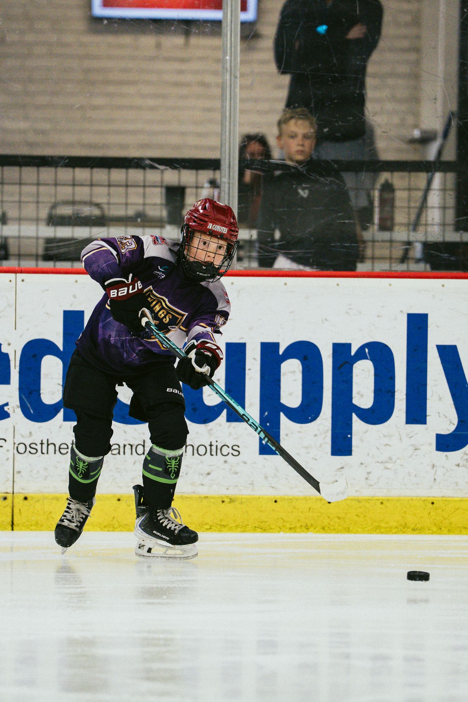 Young hockey player in purple jersey shoots puck during youth ice hockey game near rink boards.