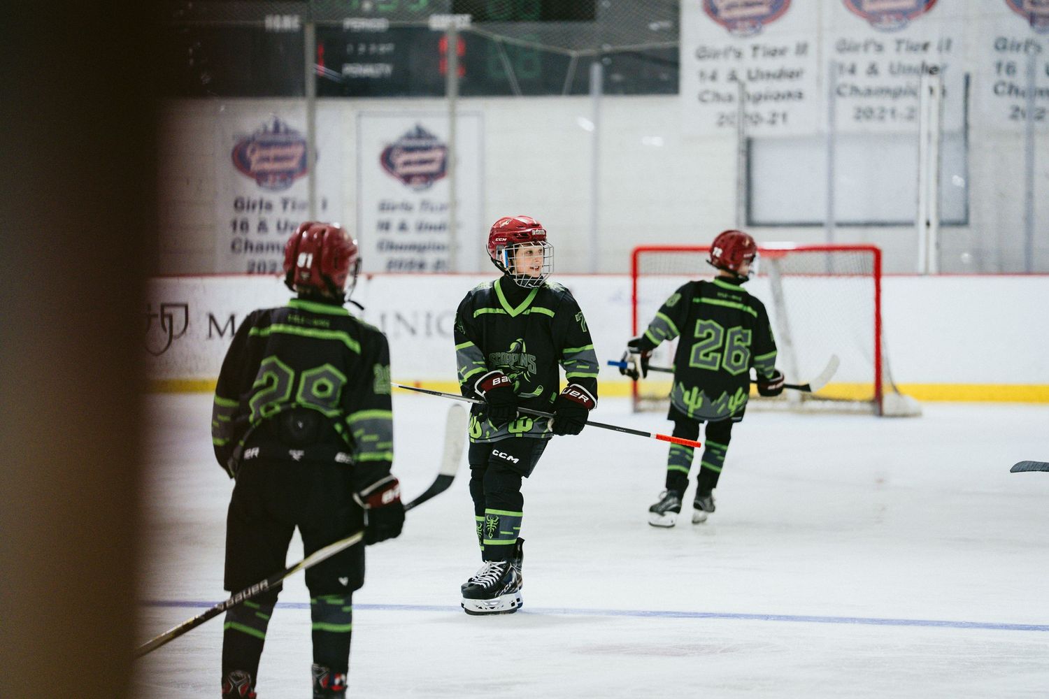 Three youth hockey players in black green uniforms stand on ice near red goal net during game break.