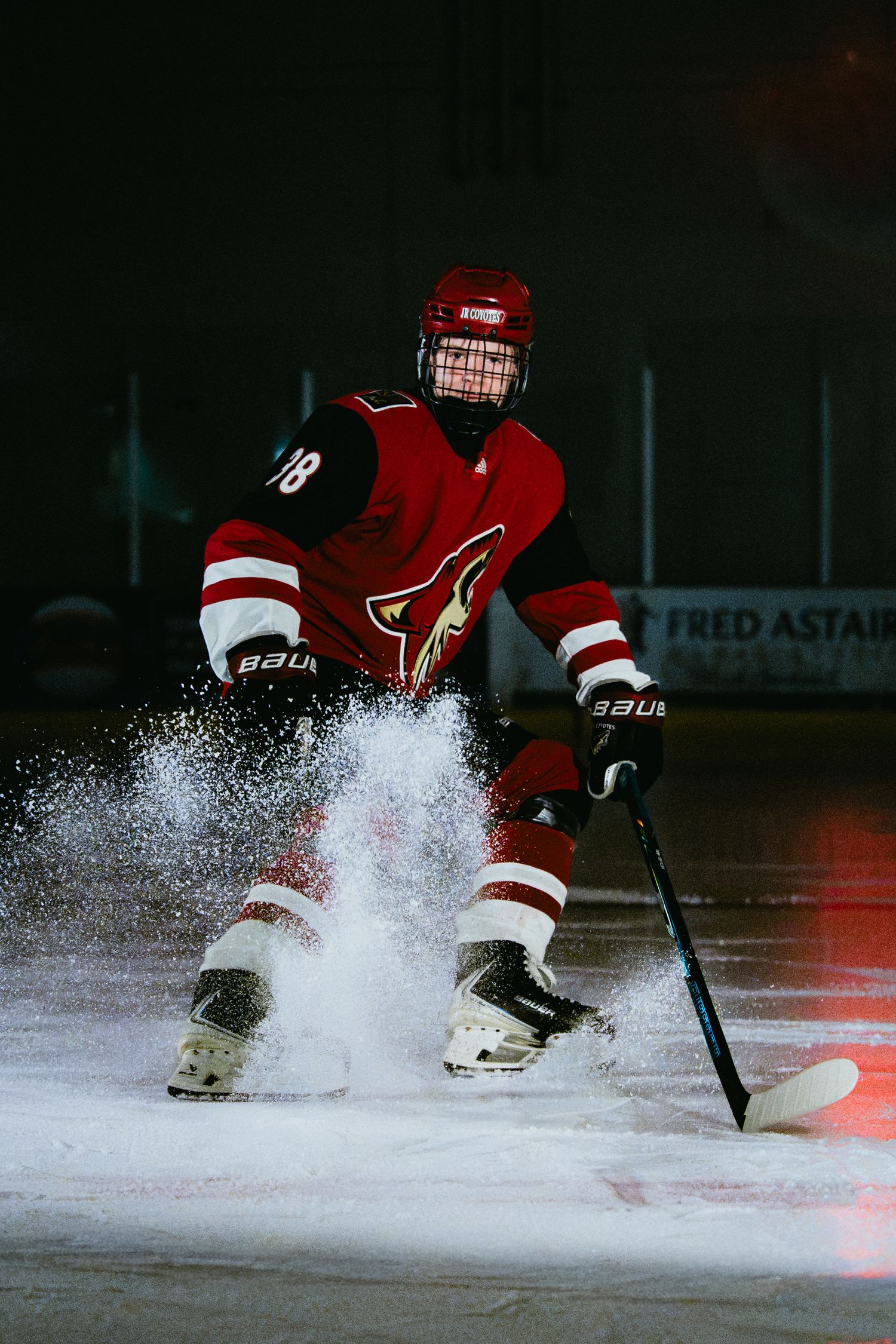 Hockey player in red Coyotes jersey stopping on ice, spraying snow, during a dramatic dark-lit rink action shot.
