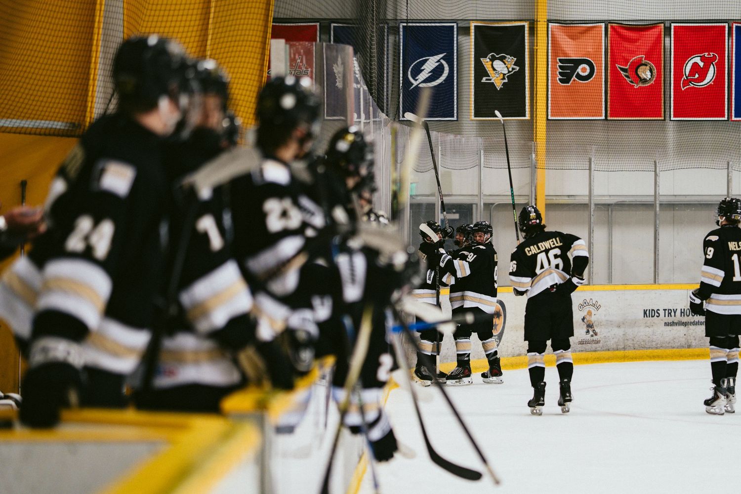 Youth hockey players in black uniforms on ice rink bench with NHL team banners displayed on arena wall.