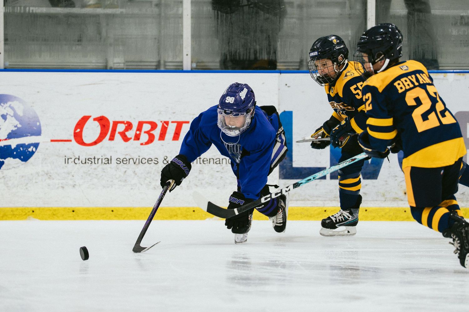 Ice hockey players compete on rink, blue team player reaches for puck against two yellow-black opponents near Orbit advertising board.