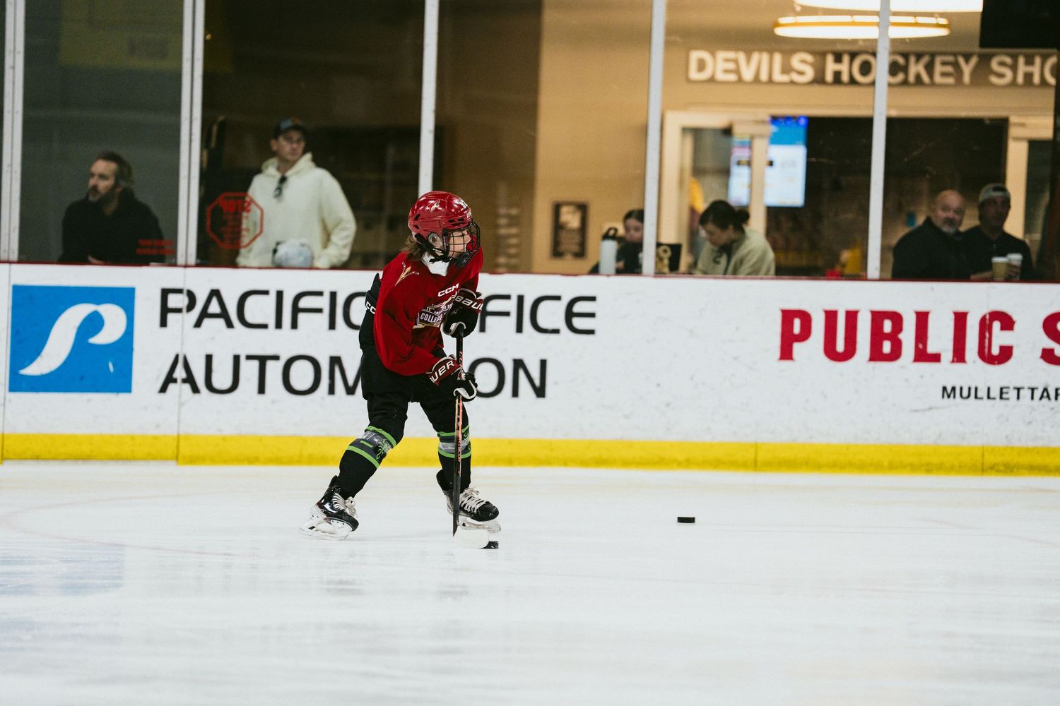 Young hockey player skating with puck at Devils Hockey Shop rink with Pacific Office Automation banner.