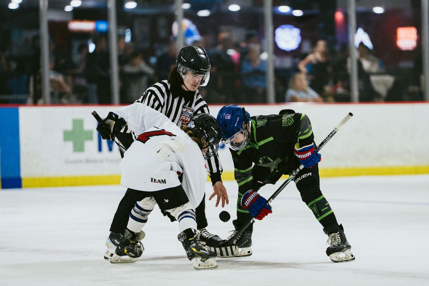 Referee oversees a face-off between two youth hockey players in an indoor ice rink.