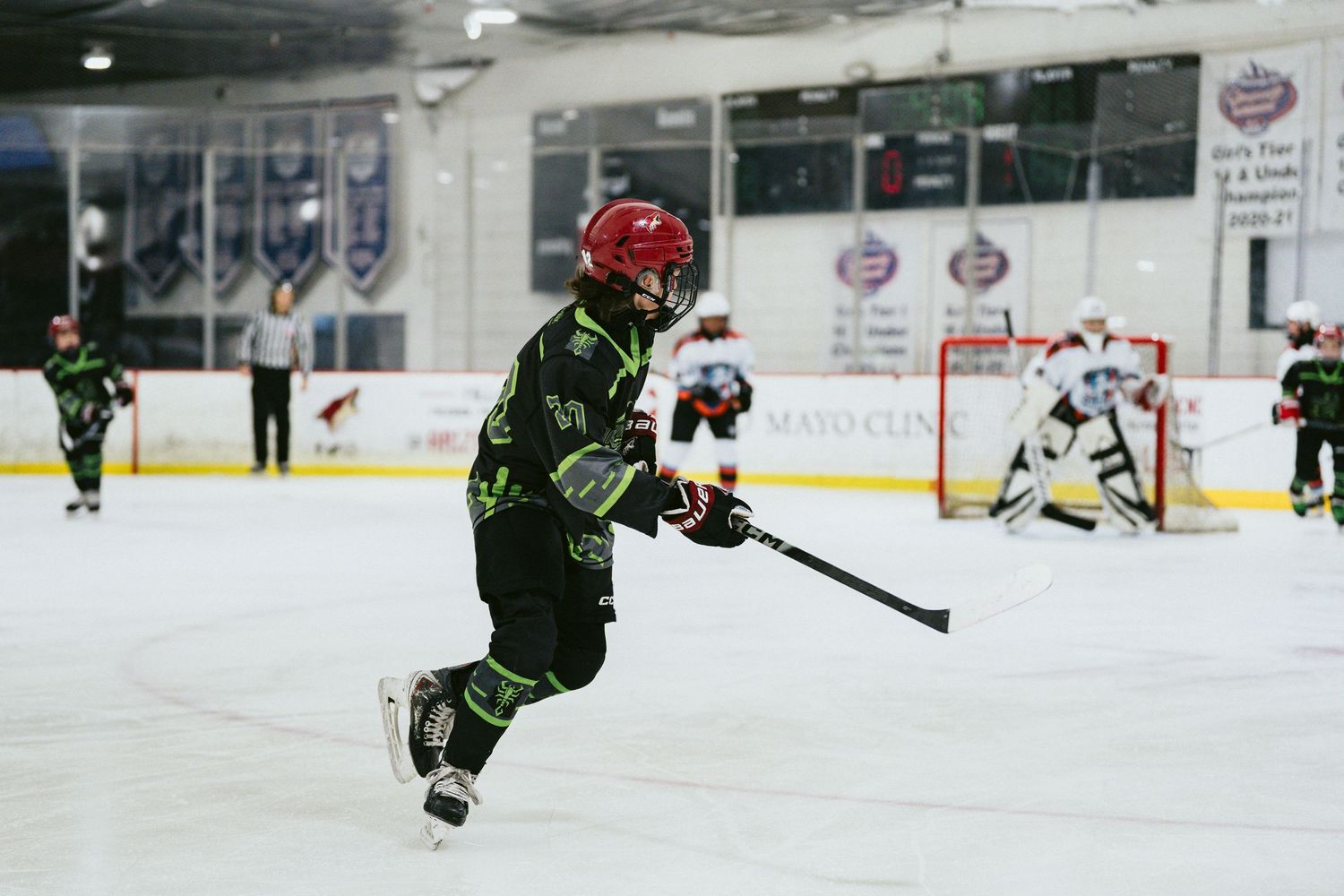 Young hockey player in black and green uniform with red helmet skating on indoor ice rink during youth hockey game.