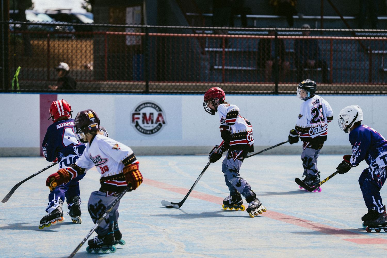 Kids play roller hockey on outdoor rink during FMA youth league game wearing colorful team jerseys.