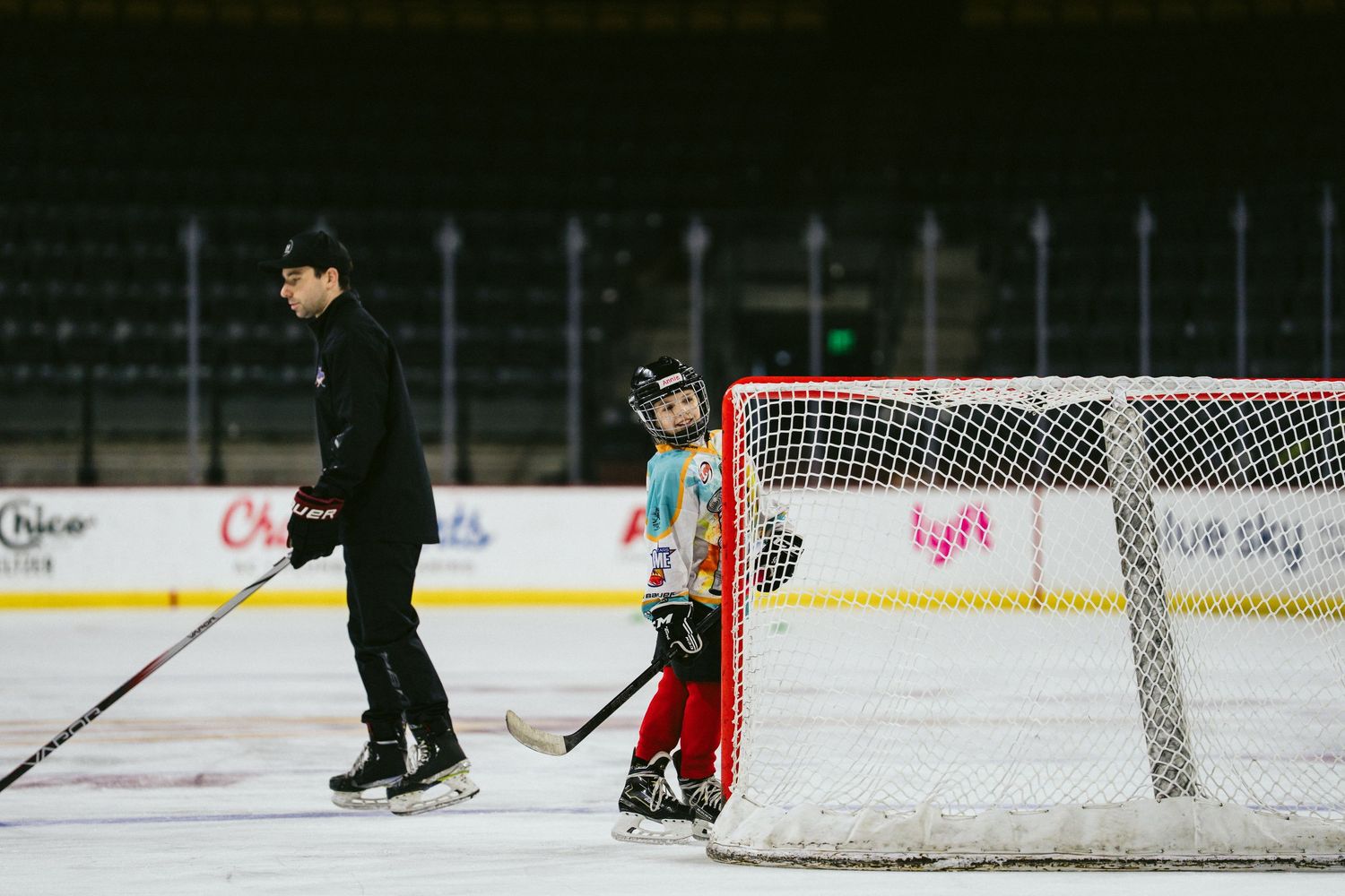 Youth hockey player in red gear standing by goal net with coach during ice practice.