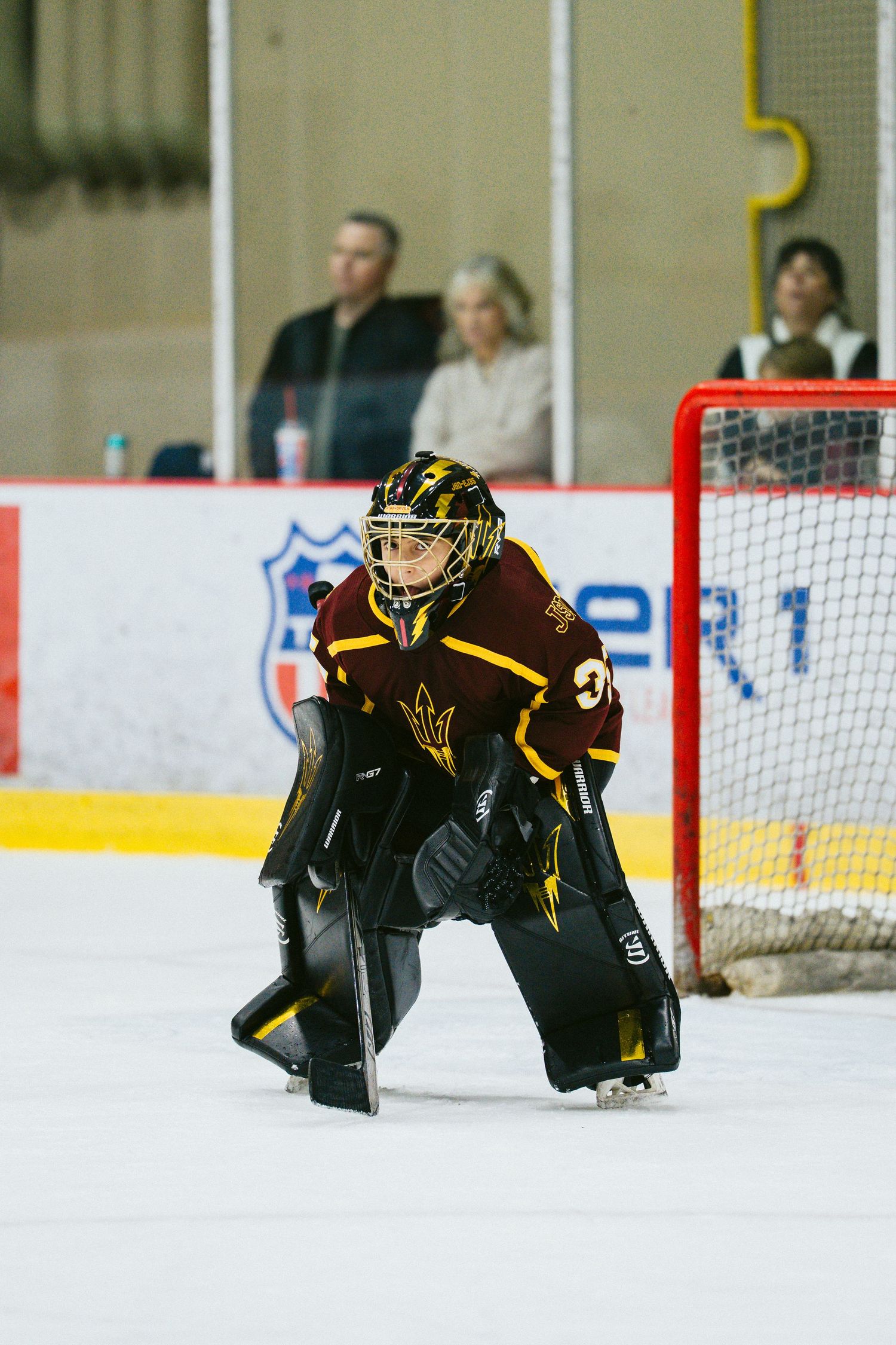 Hockey goalie in maroon and gold uniform crouches in ready position on ice during game.