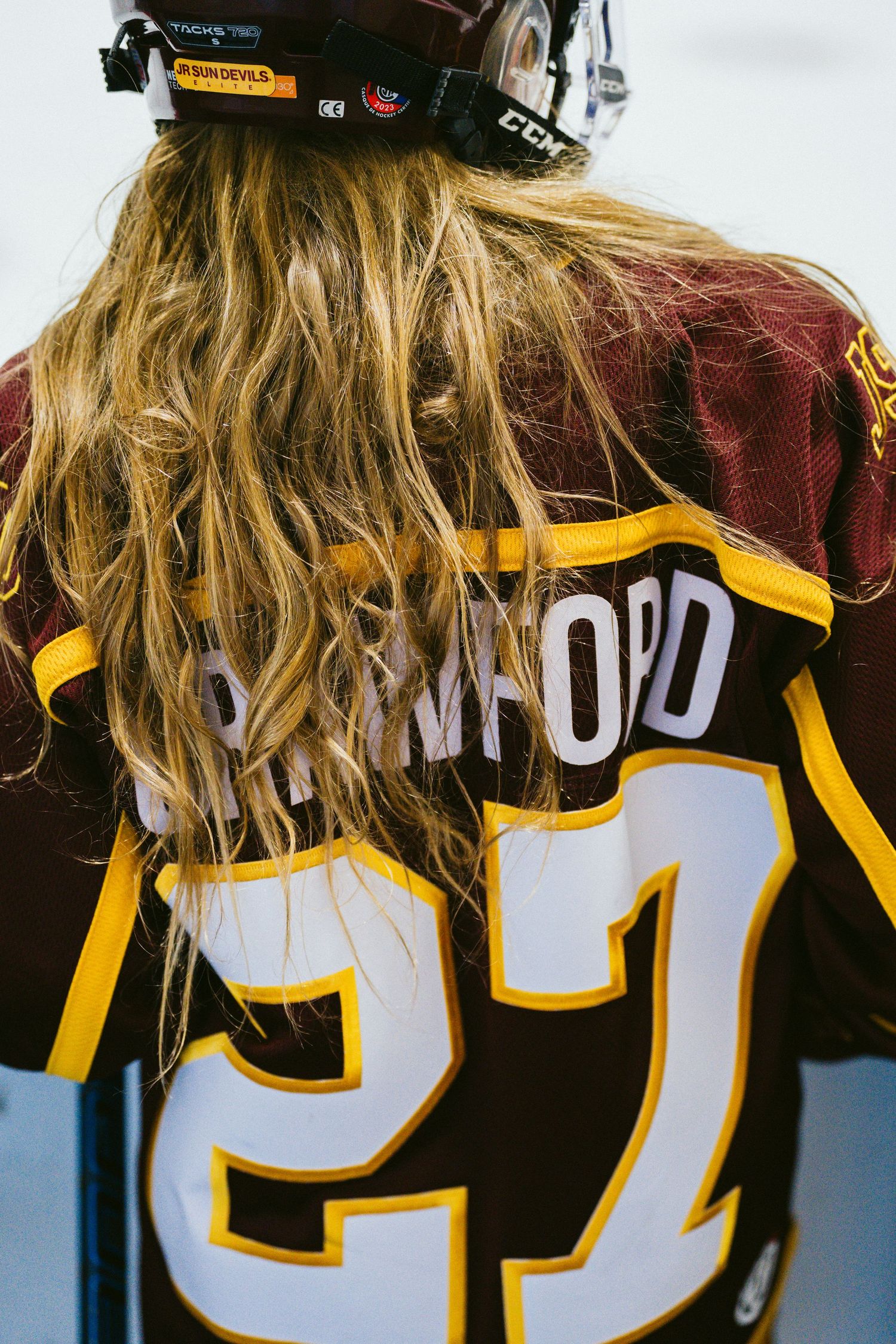 Close-up view of hockey player wearing maroon and gold jersey number 27 with long blonde hair.