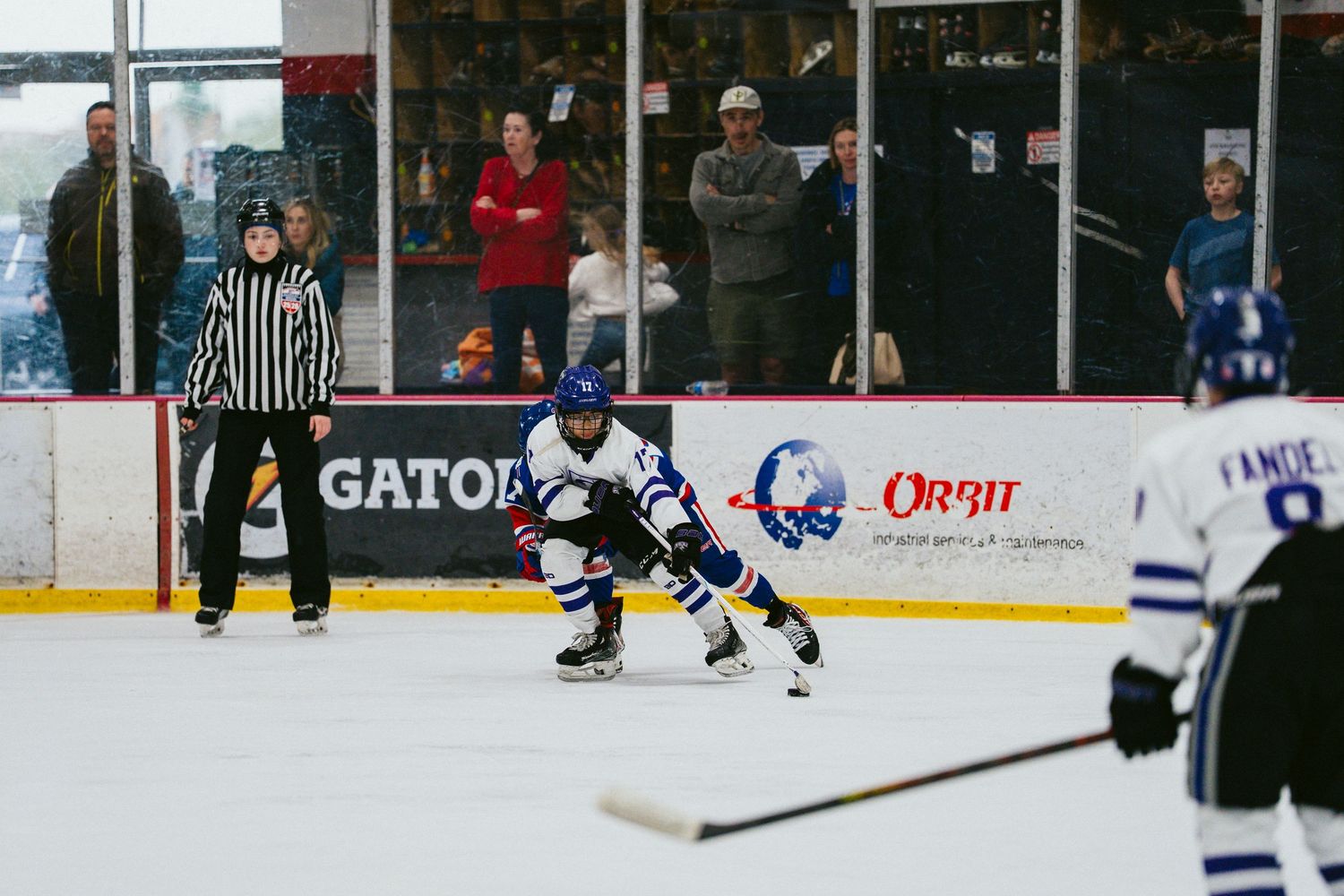 Youth hockey game in progress with referee and spectators watching from sidelines.