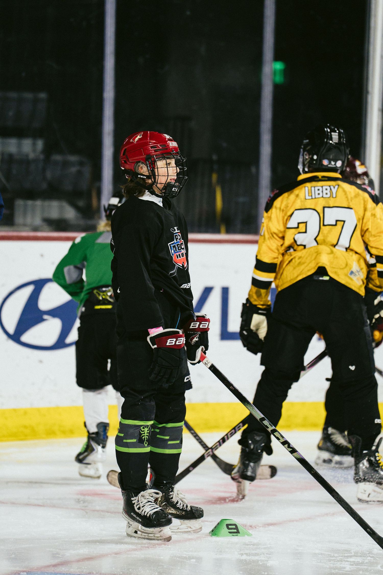 Young ice hockey player in red helmet and black uniform stands on rink near player number 37 Libby.