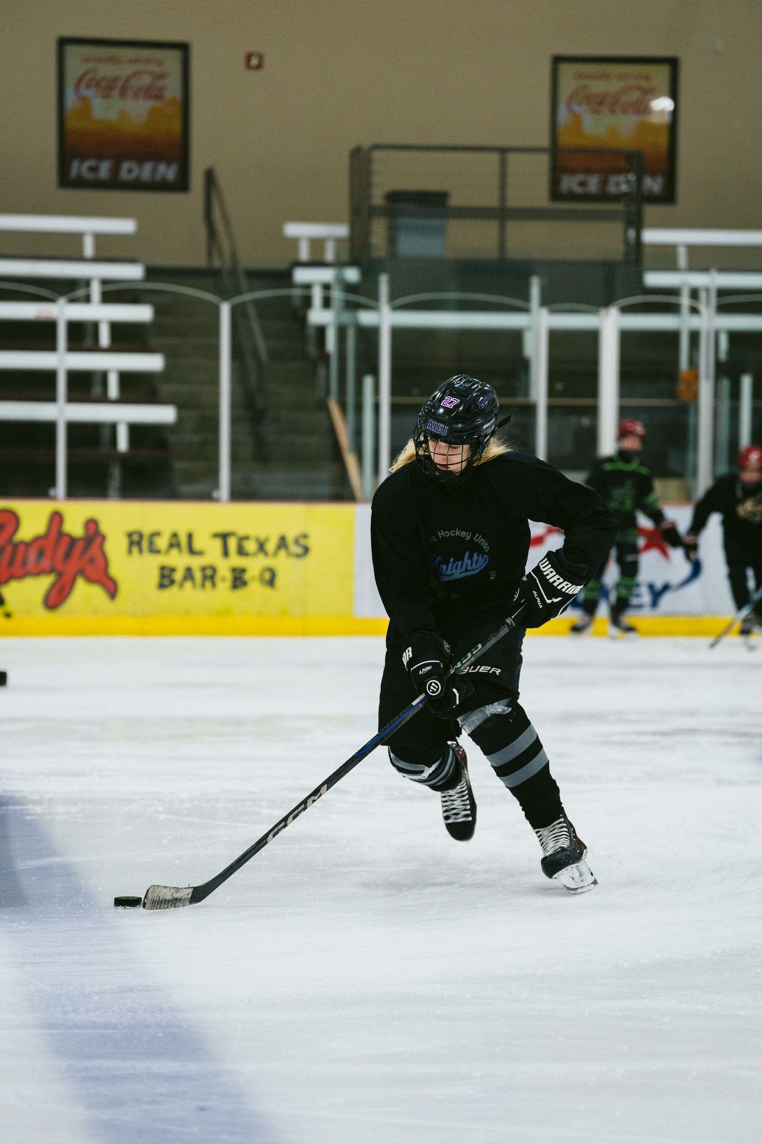 Female hockey player in black uniform skating with puck during ice hockey practice at indoor rink.