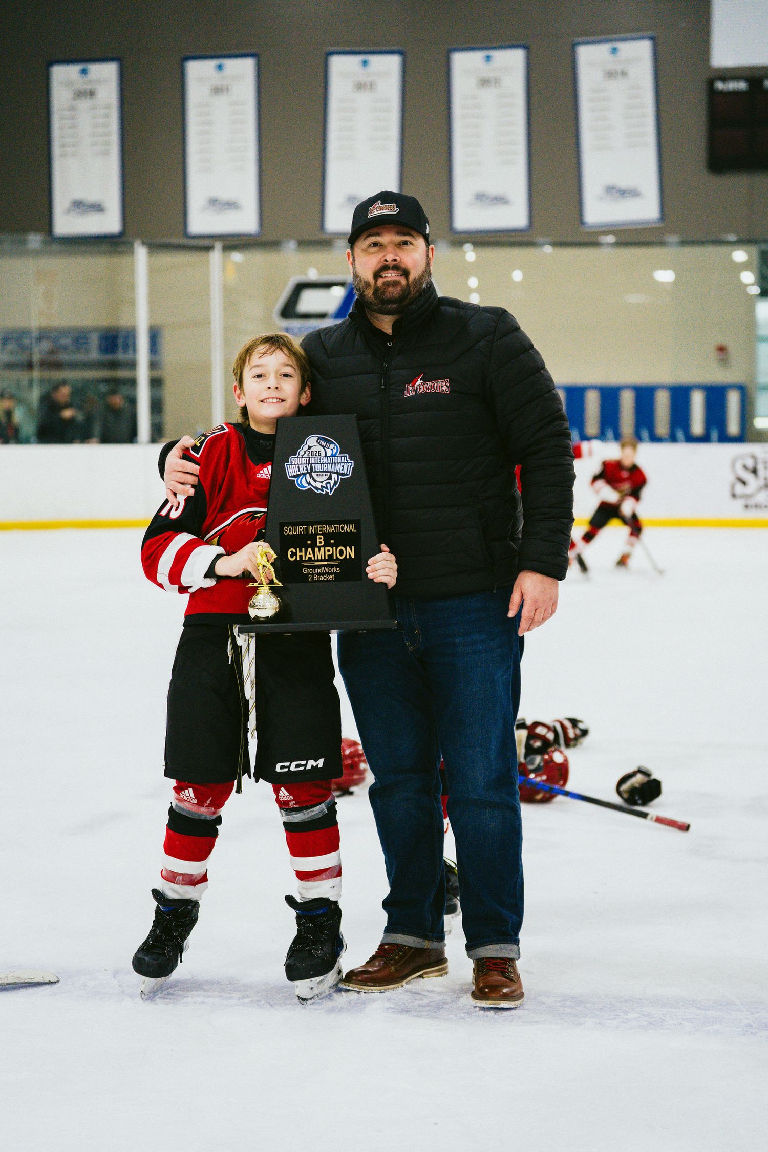 Youth hockey player poses with adult on ice rink while holding championship plaque.