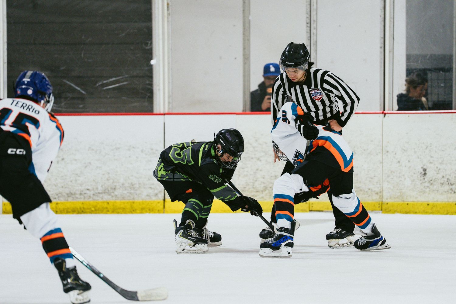Youth ice hockey face-off between two players as referee drops puck during indoor rink game.