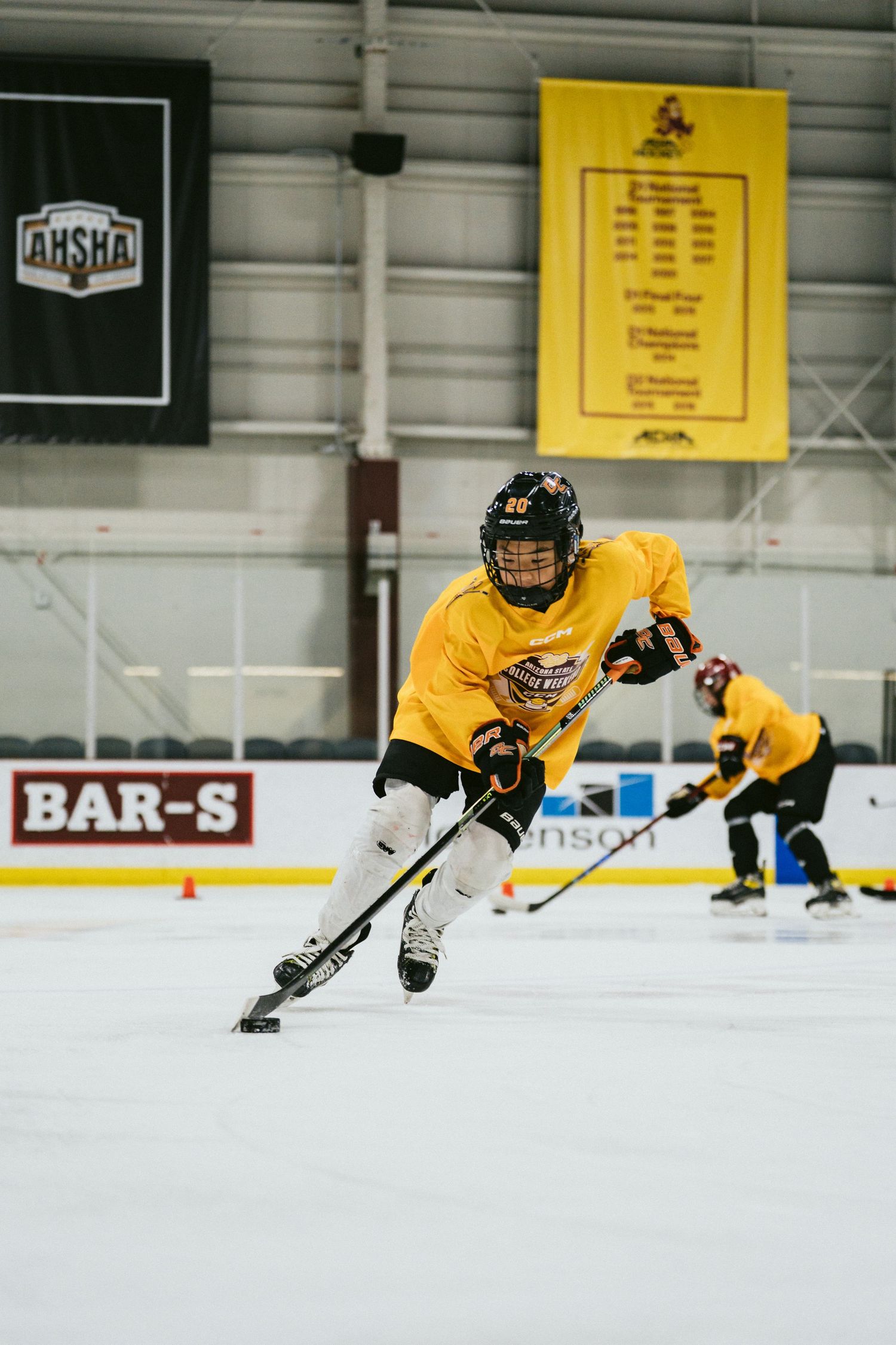 Young hockey player in yellow jersey dribbles puck across ice during indoor training session with AHSHA banner.