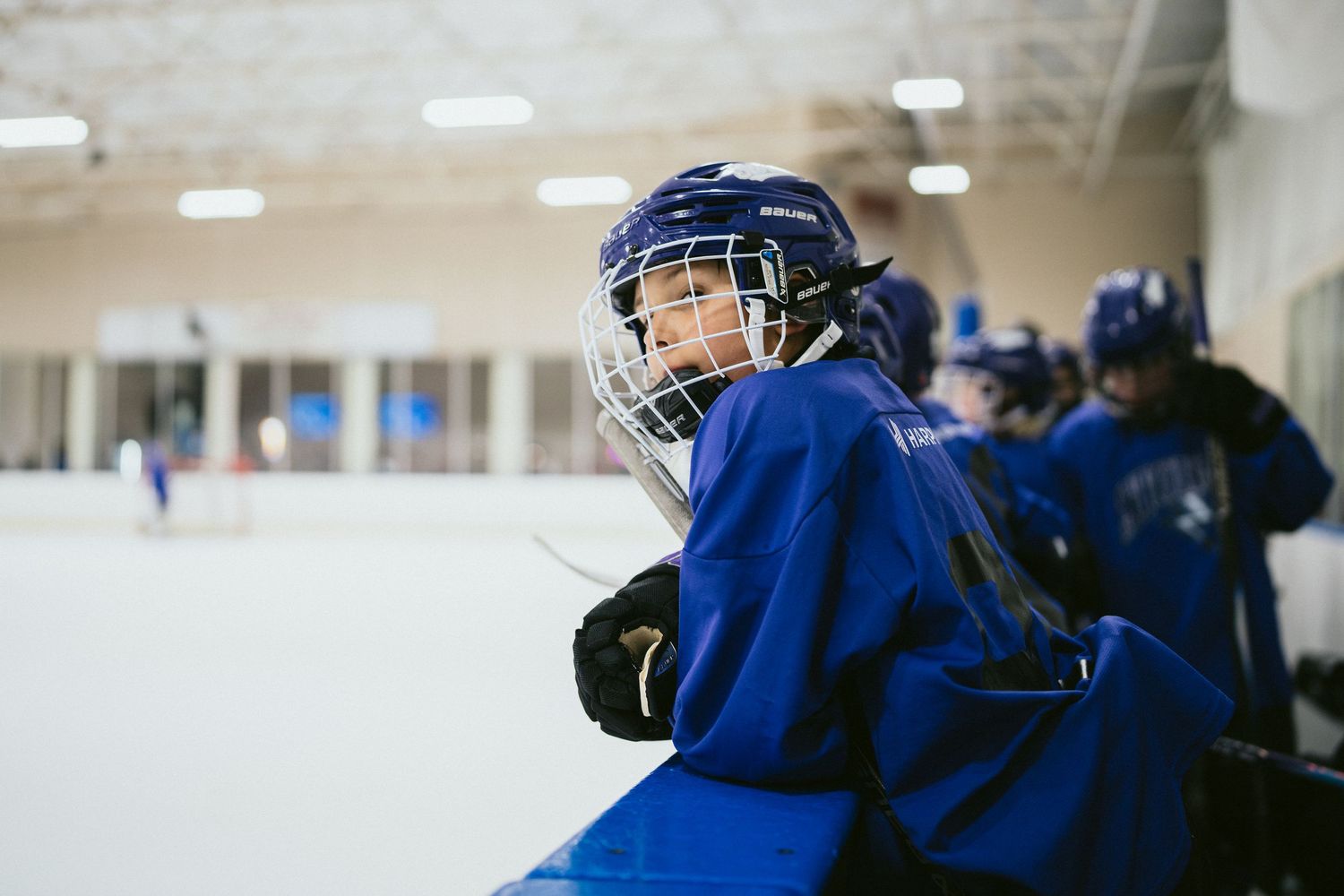 Young hockey player in blue jersey and helmet leaning on rink boards during practice session.