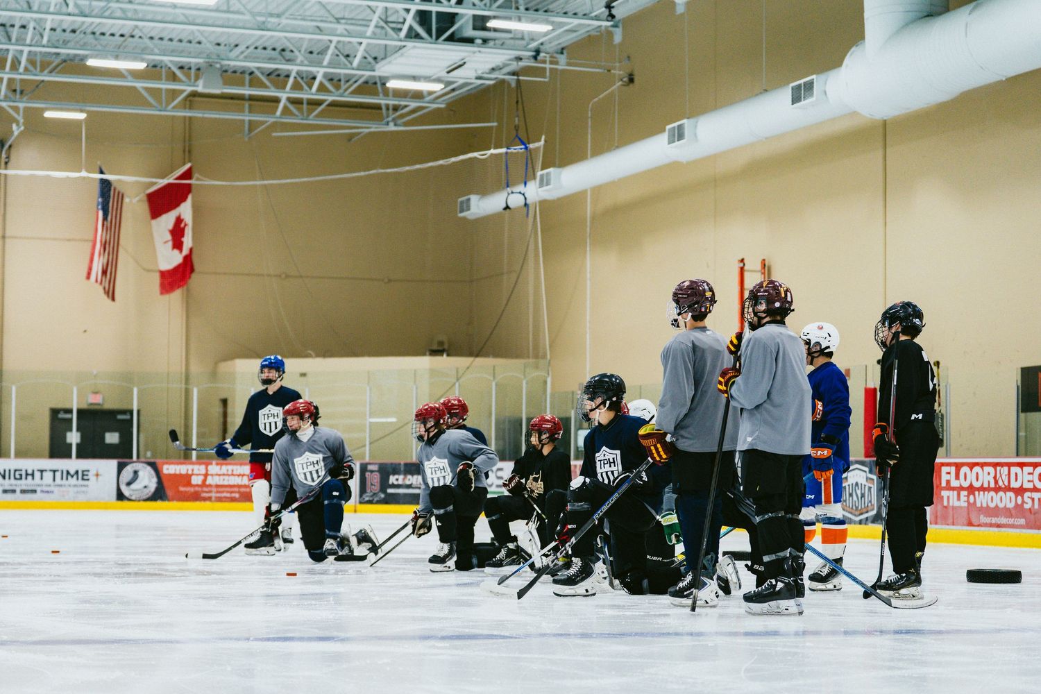 Hockey team listens to coach during indoor ice rink practice near Canadian and American flags.