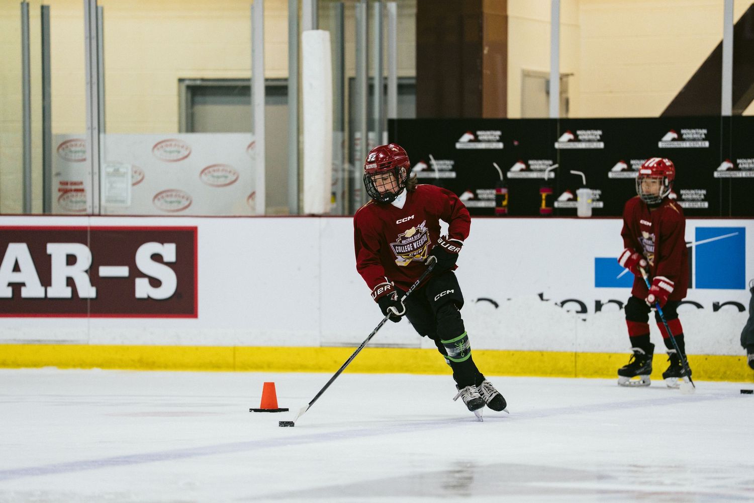 Young hockey players in red uniforms practice skating drills with orange cones on an indoor ice rink.