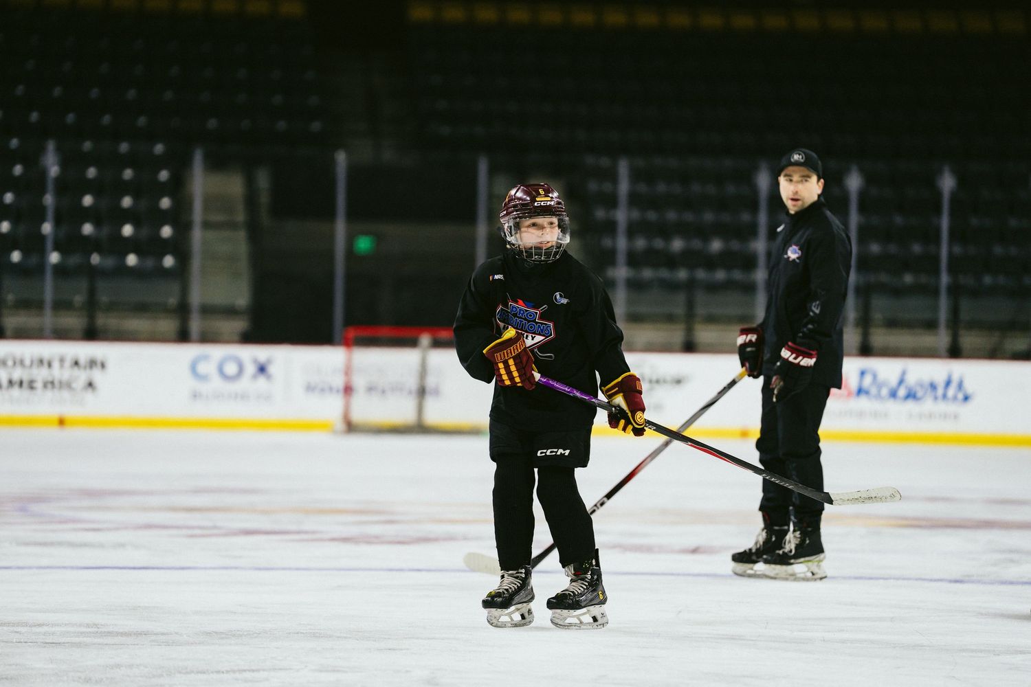 Young hockey player stands on ice with coach during youth hockey training session at indoor rink.