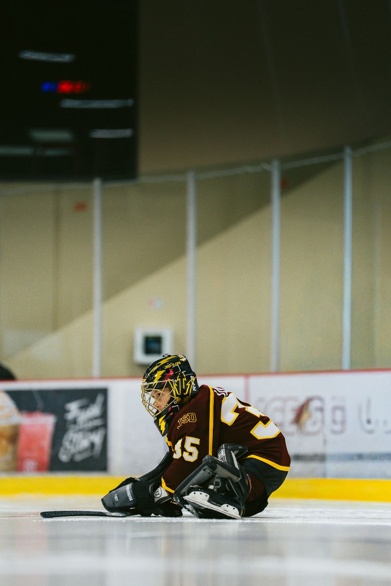 Hockey goalie in black and gold uniform sits on ice during practice session at indoor rink.