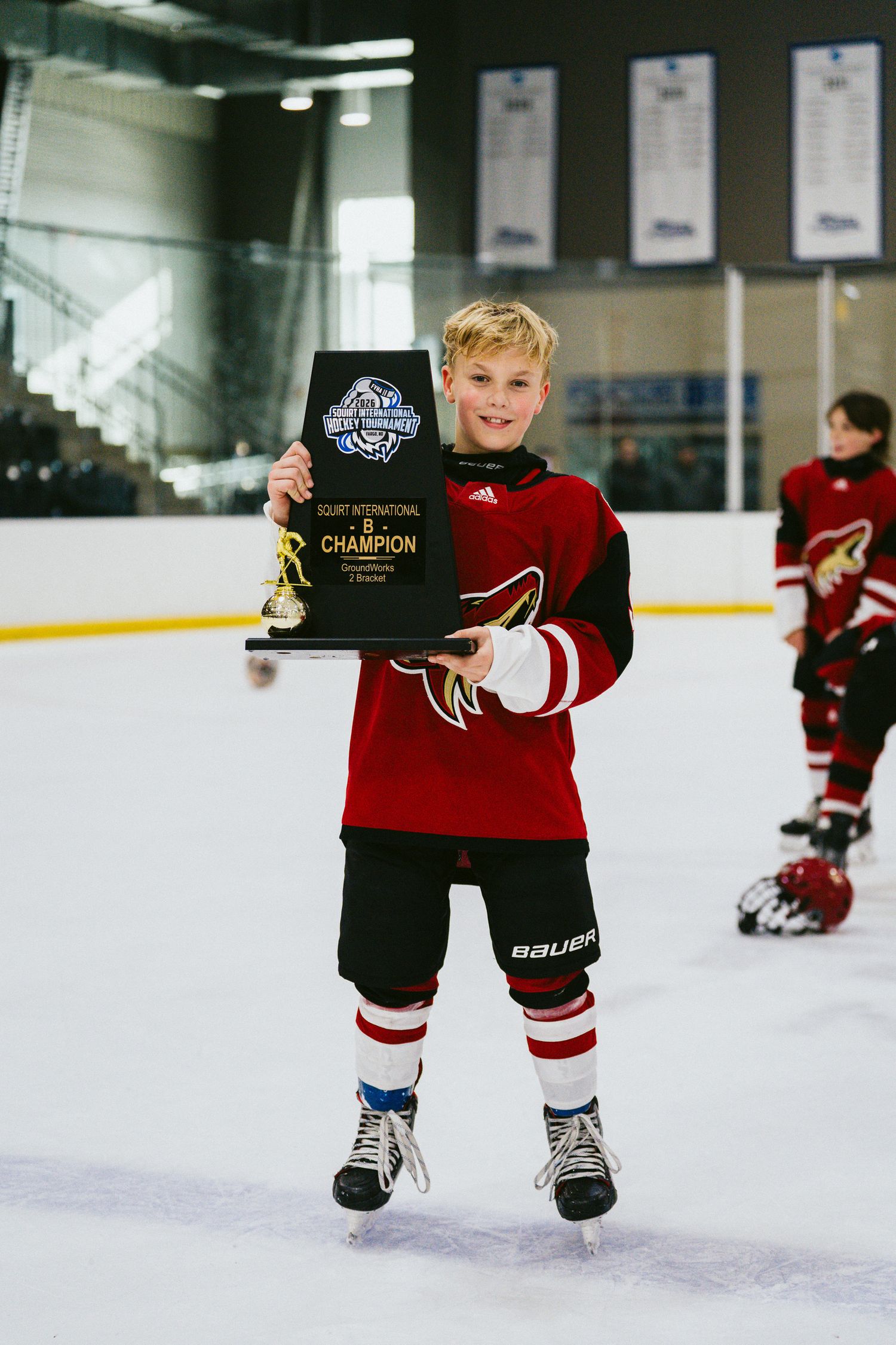 Young hockey player in red jersey and black gear proudly displays award on ice rink.