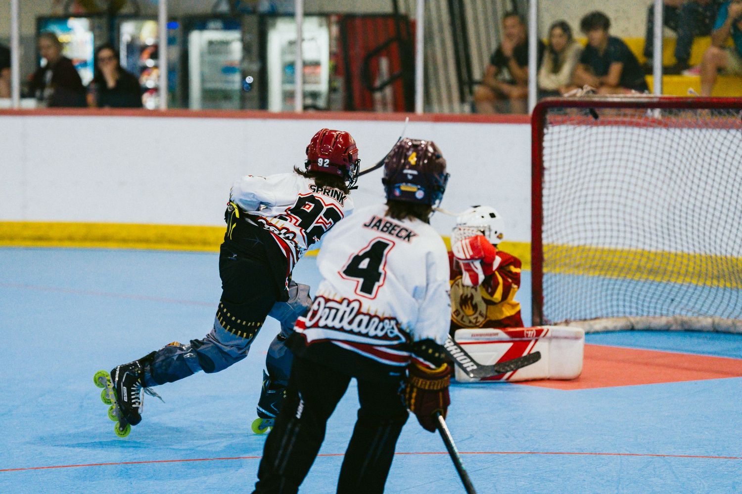 Youth roller hockey players battle near the goal as player number 4 in Outlaws jersey attempts a shot.