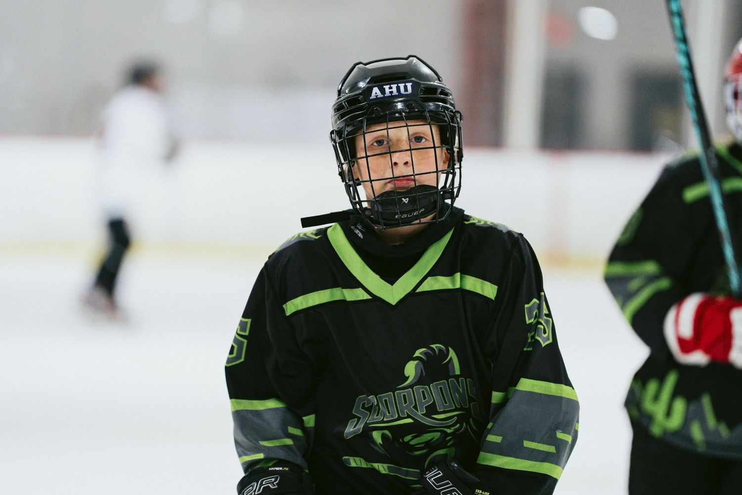 Close-up of young hockey player wearing AHU helmet and black green Scorpions jersey on indoor ice rink.