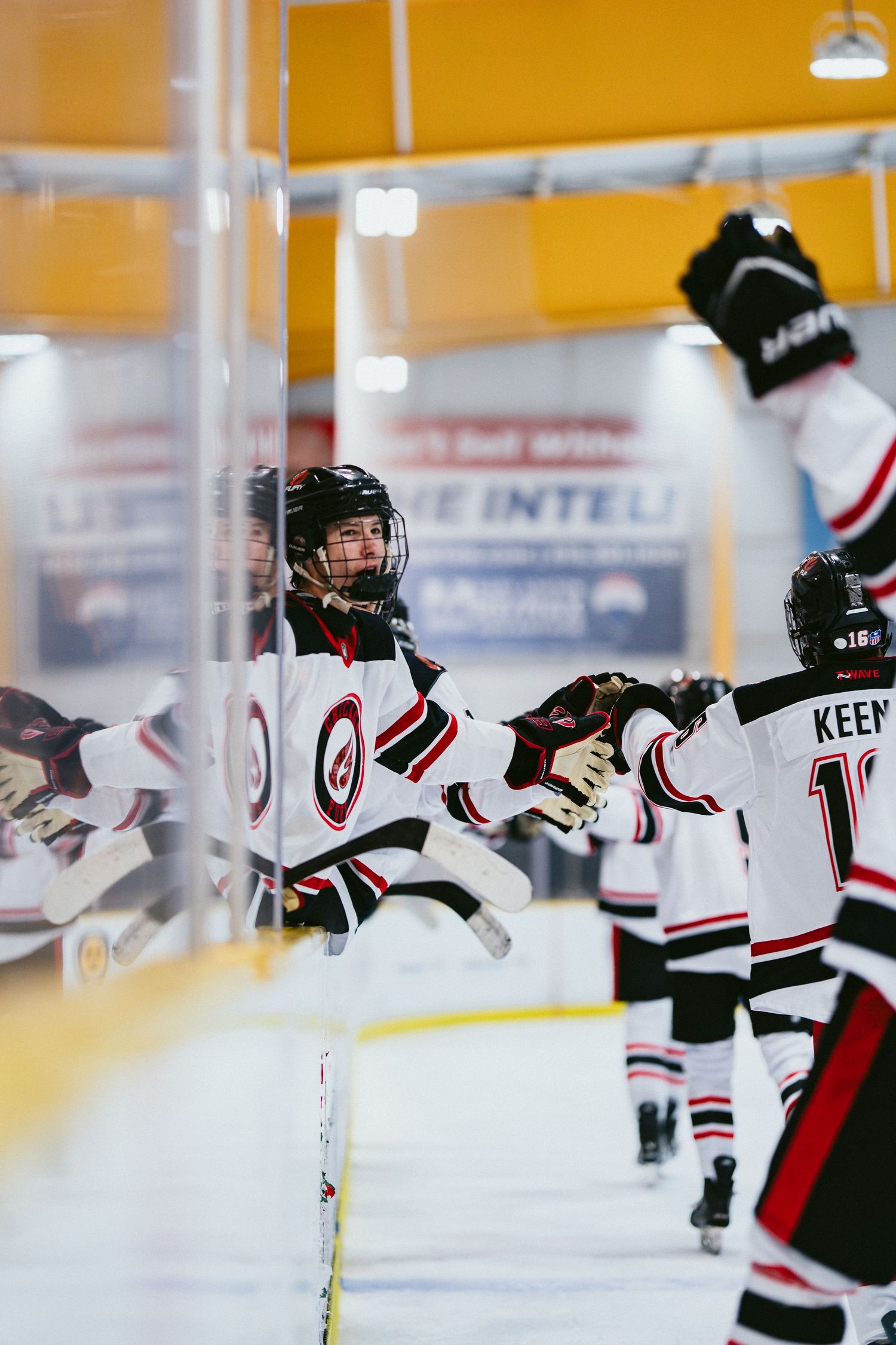 Youth hockey players in white and black uniforms celebrate a goal by high-fiving along the boards.