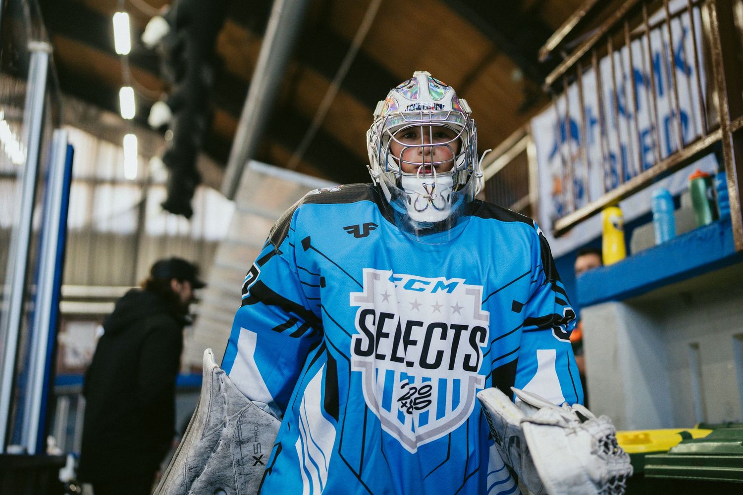 Hockey goalie in blue CCM Selects uniform and mask walks through arena tunnel ready for game.