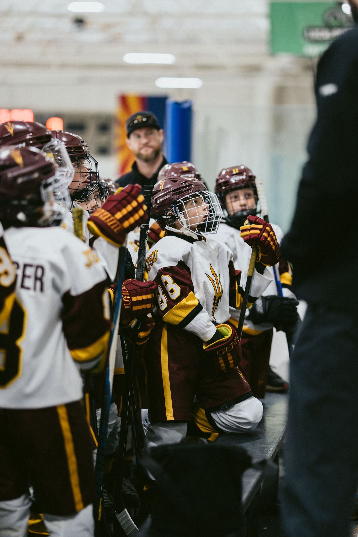 Young hockey players in maroon and gold uniforms kneel and listen to their coach during a game.