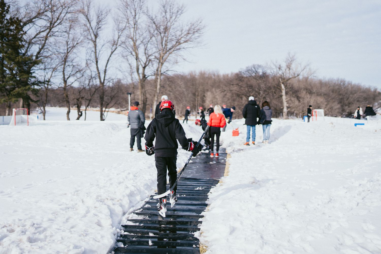 People walking on a wooden boardwalk through snowy winter landscape with bare trees.