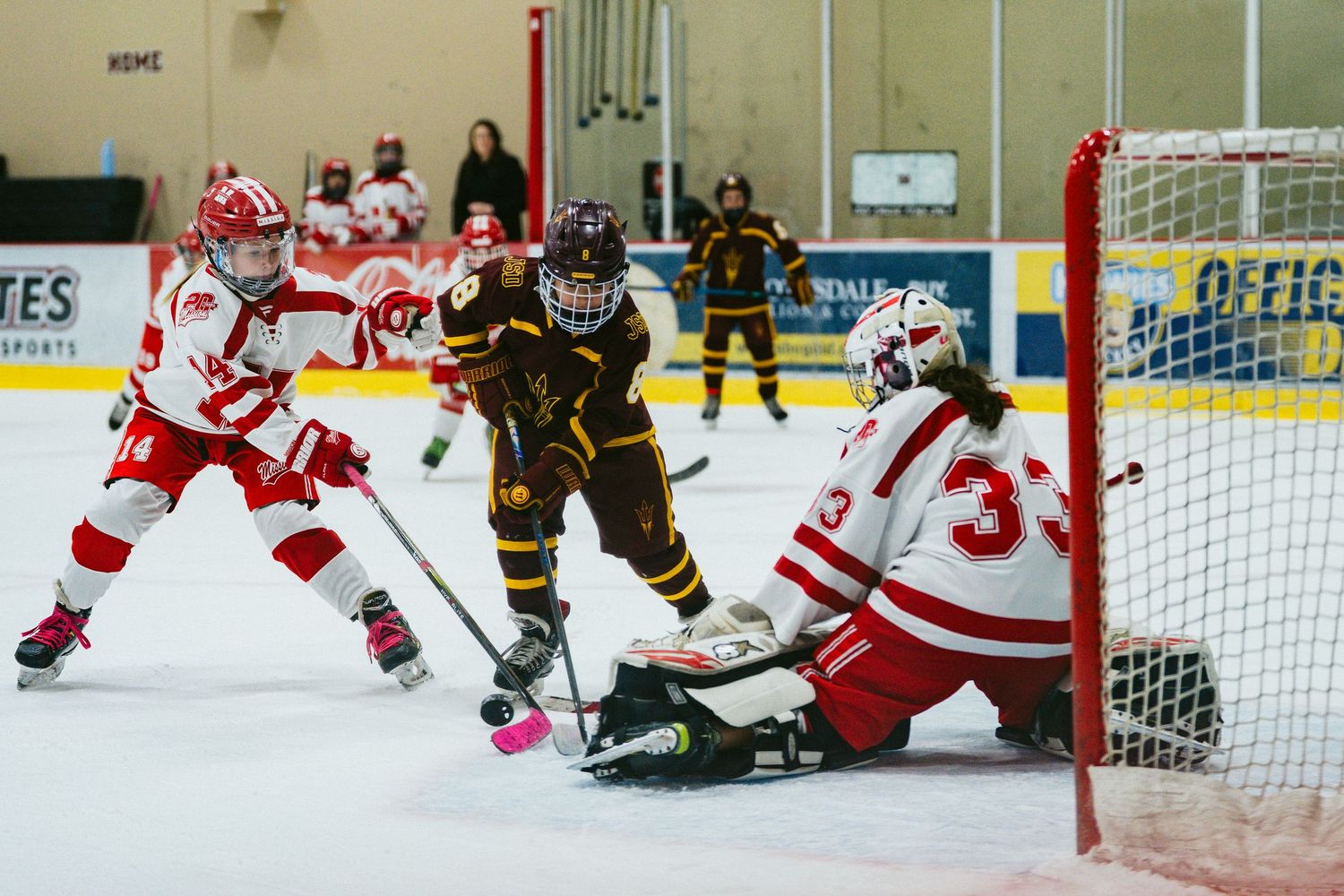 Ice hockey game action shows players from opposing teams in red and brown uniforms near the goal.