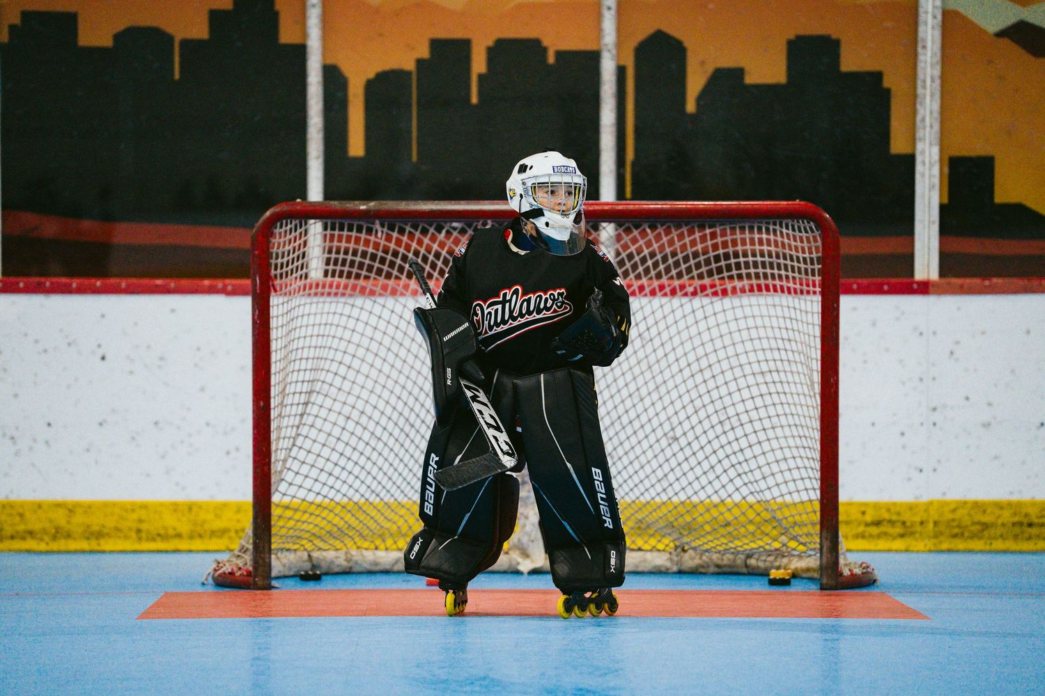 Young goalie in black gear guards the net during an indoor roller hockey game on a blue rink.