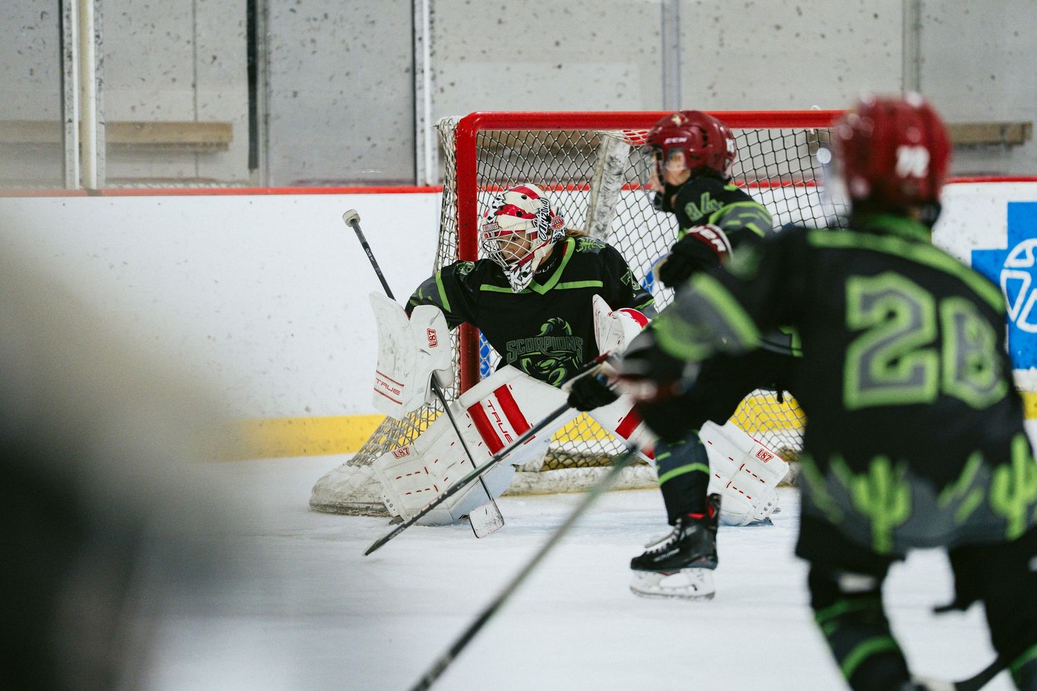 Youth hockey goalie in black gear defending red net during indoor ice hockey game.