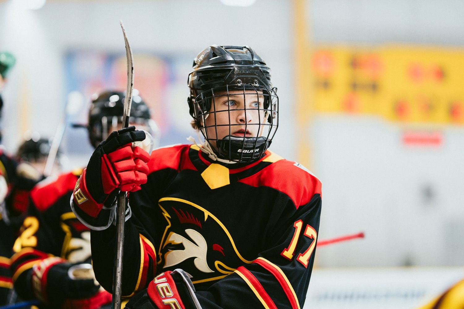 Youth hockey player in red and black jersey raises their stick on the ice during an indoor hockey game.