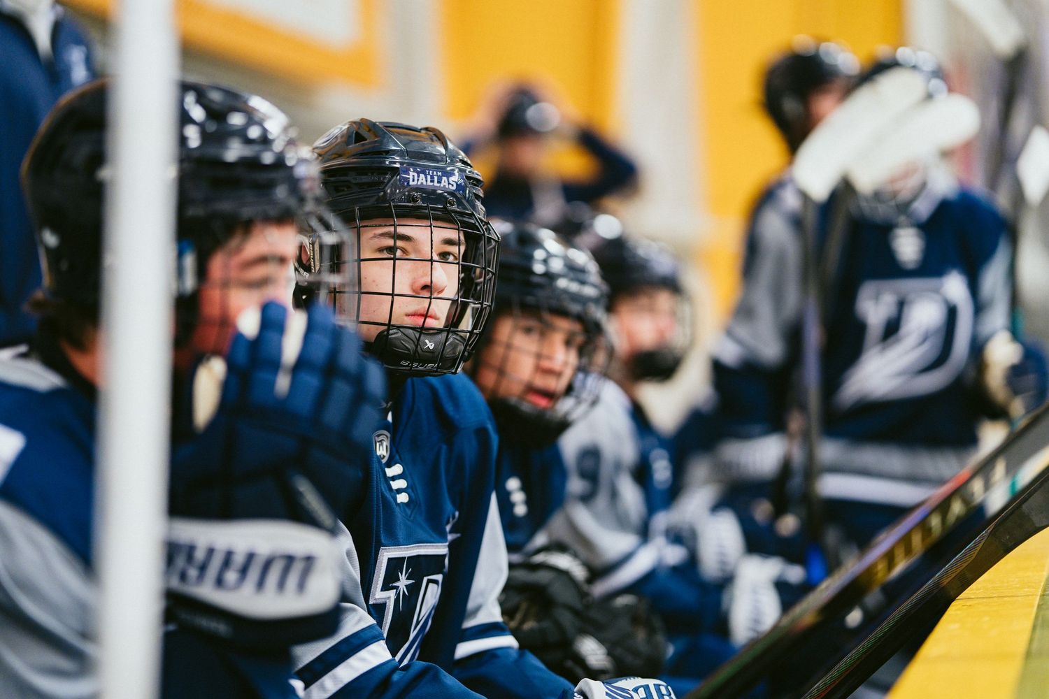 Young youth hockey players sit on the bench wearing navy blue helmets and jerseys during an ice hockey game.