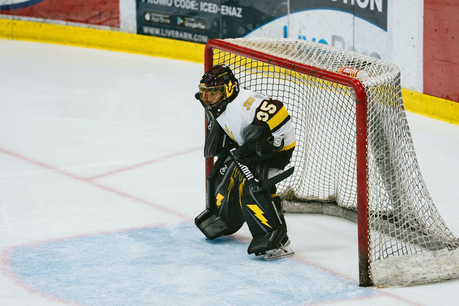 Hockey goalie in black and gold uniform guards the net during an indoor ice hockey game.