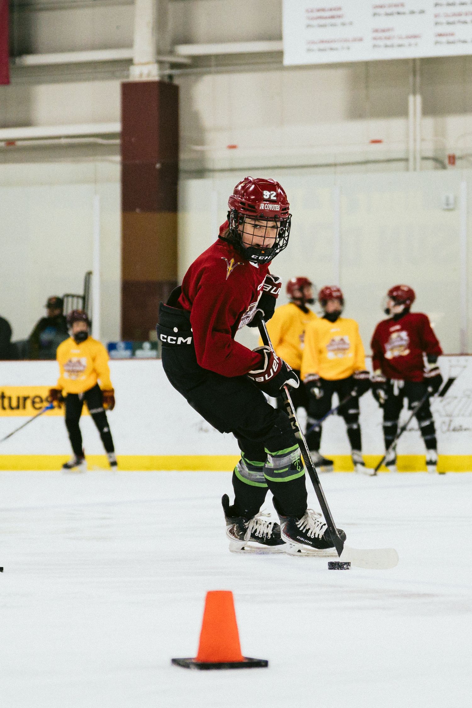 Young hockey player in red CCM jersey maneuvering puck around orange cone during indoor ice practice.