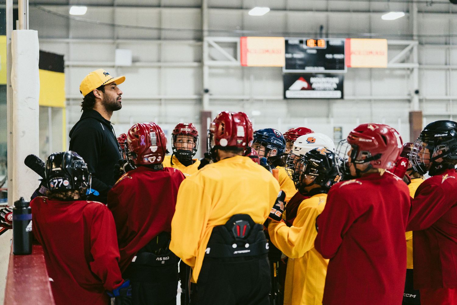Hockey coach in yellow jersey addresses a group of young players in red helmets and jerseys during an indoor rink practice.