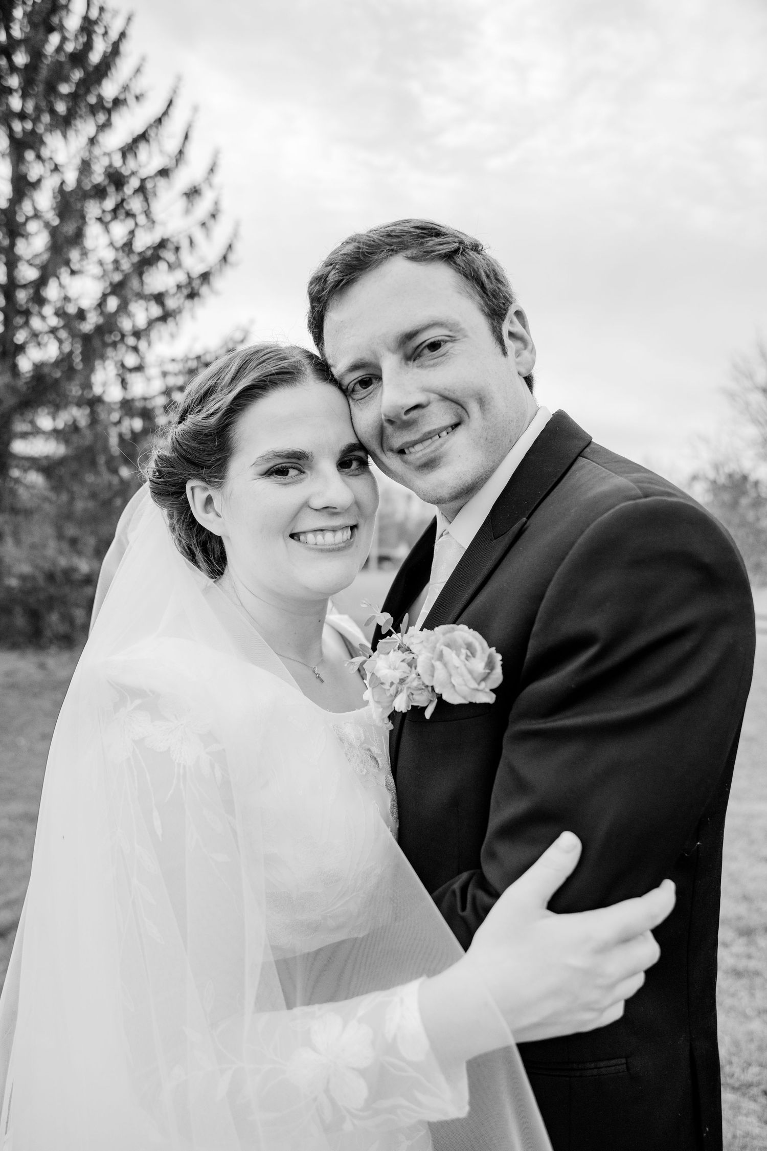 Black and white wedding portrait of smiling bride in veil and groom in suit with boutonniere embracing outdoors.