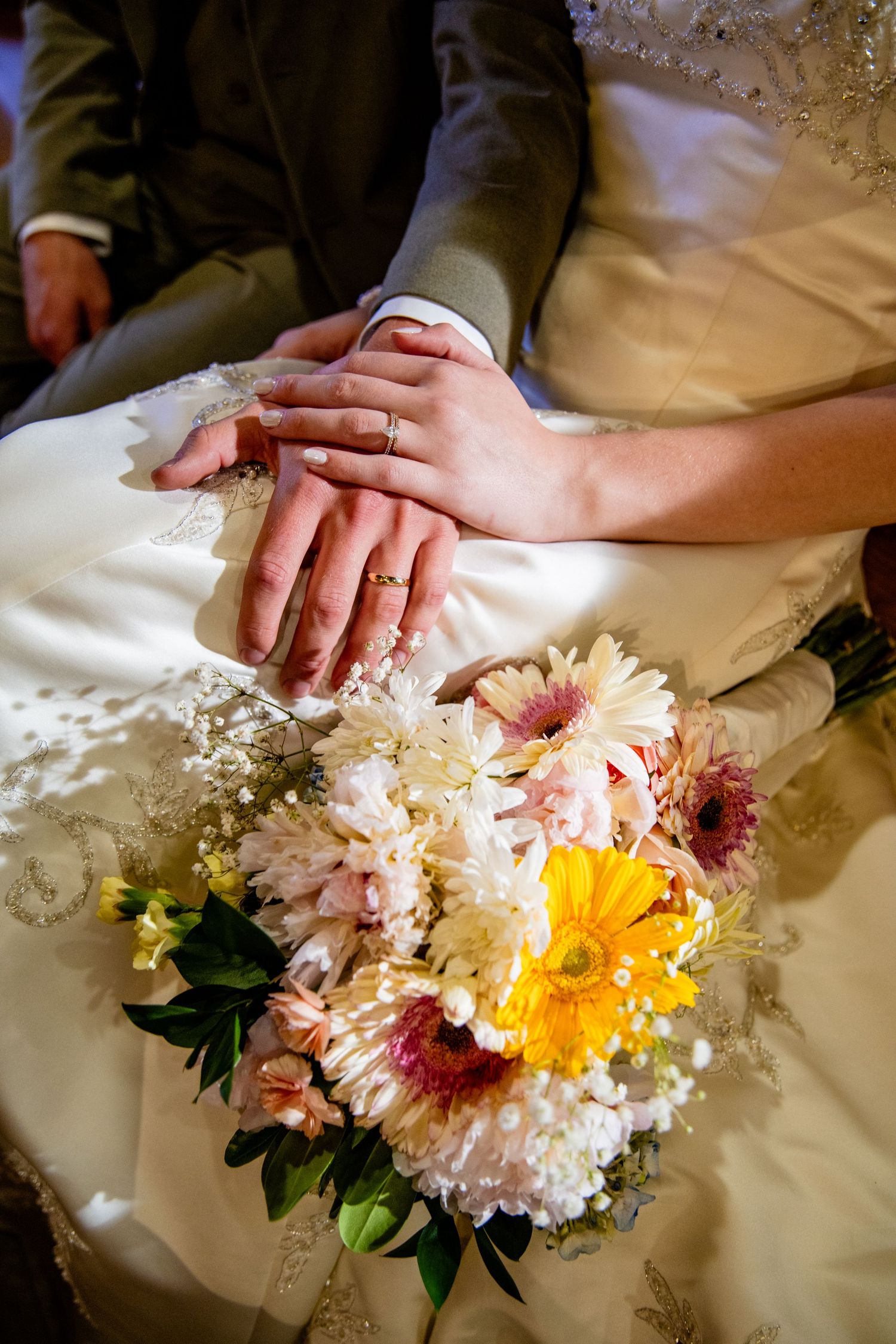 Bride and groom holding hands with wedding rings, resting on white dress beside a colorful bridal bouquet of gerberas.
