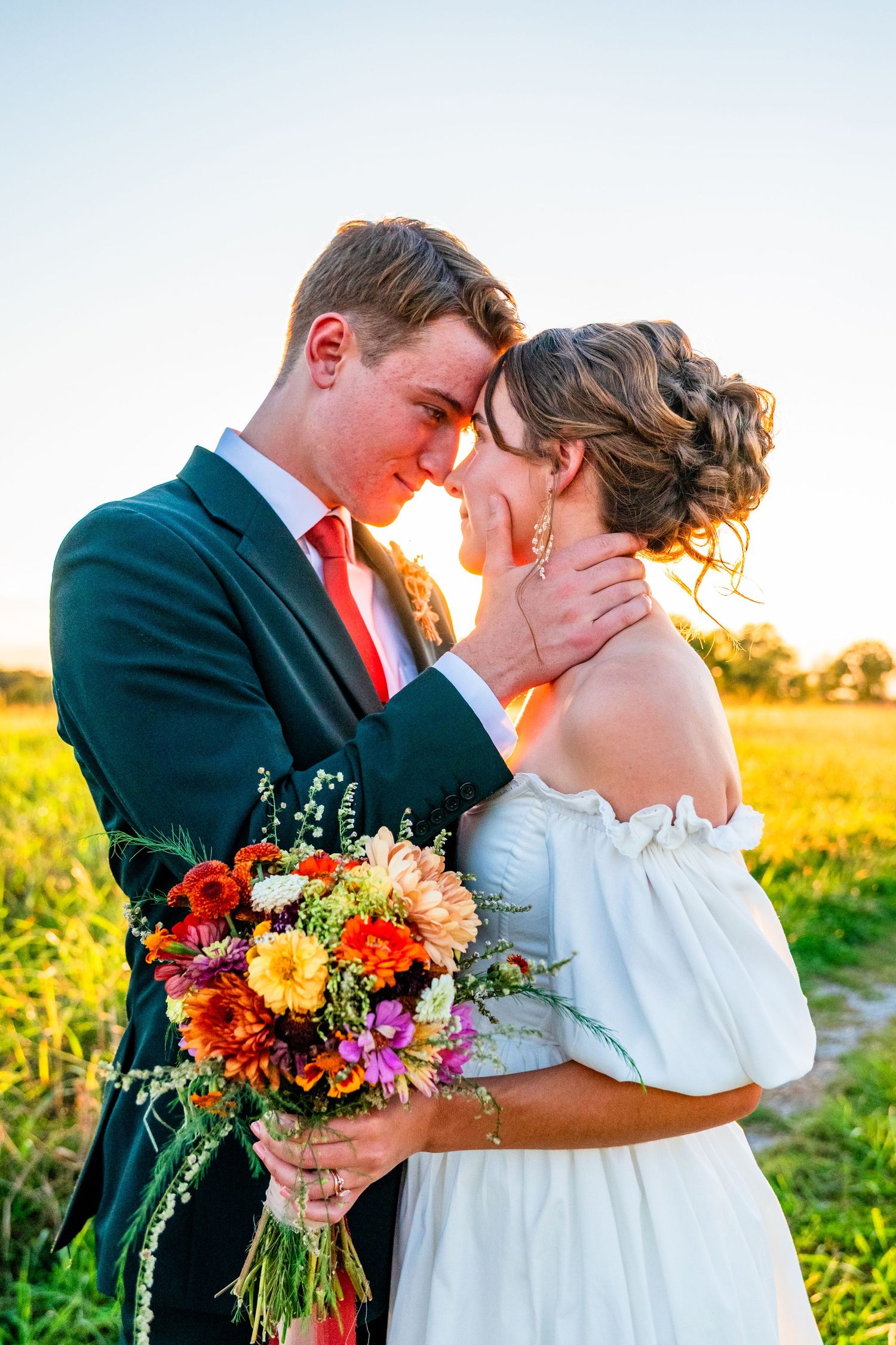 Bride and groom touching foreheads in golden sunset light, holding colorful wildflower bouquet in green field.