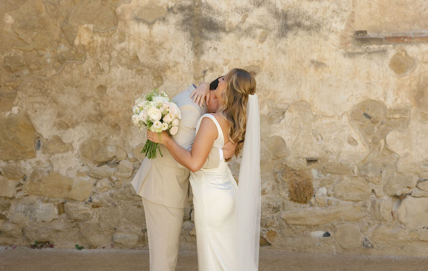 Two people in wedding attire share an intimate moment while holding a white floral bouquet against a rustic stone wall.