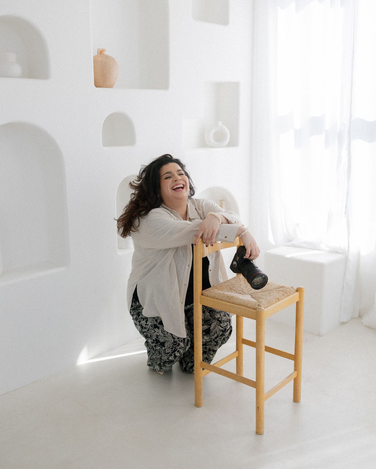 Person in casual attire laughing while sitting on wooden stool in bright white minimalist room.