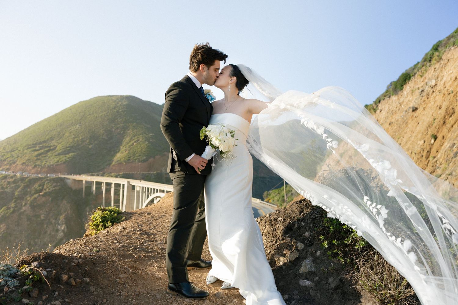 Couple kissing in wedding attire on coastal cliffside, bride's veil flowing in wind with mountain bridge backdrop.