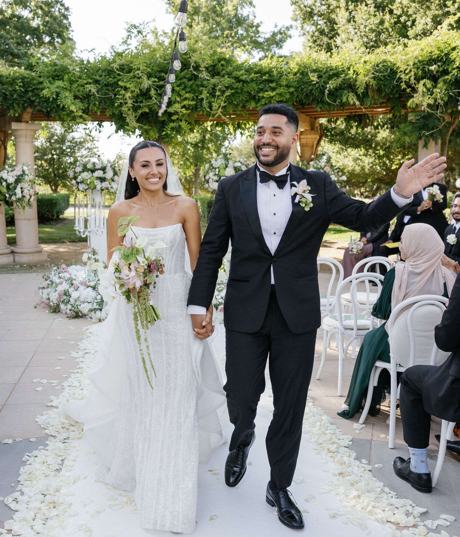 A smiling bride and groom walk down the aisle at an outdoor wedding ceremony surrounded by white floral petals.